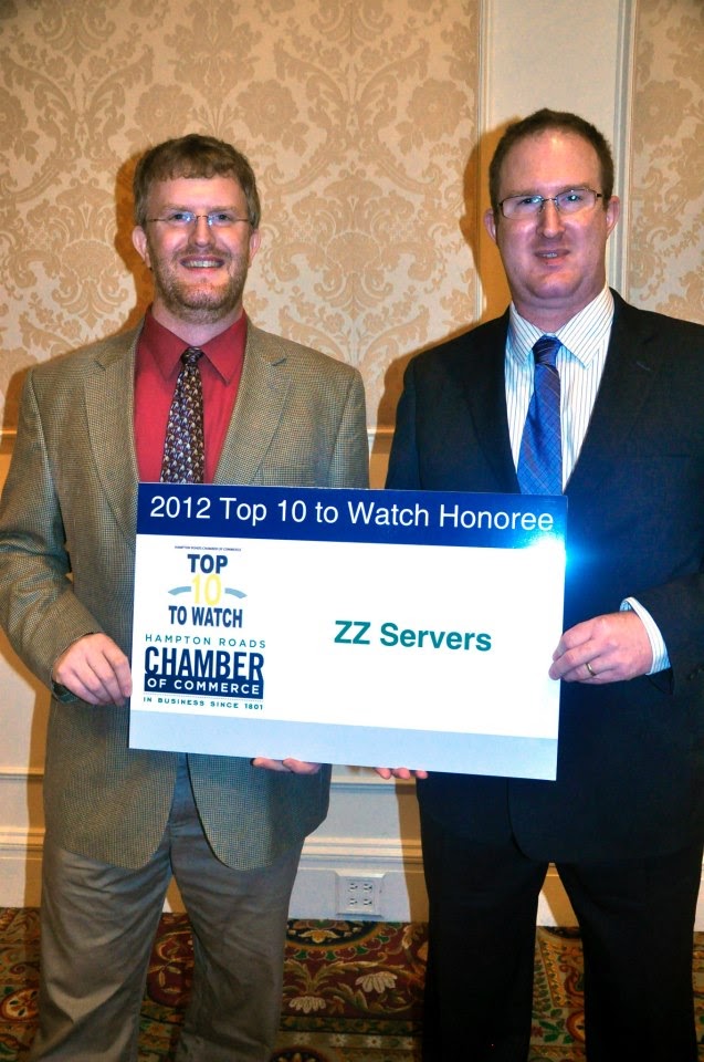 Two men in suits holding a "2012 top 10 to watch honoree" sign from the chamber of commerce, smiling at a formal event. Modified description: two gentlemen in suits