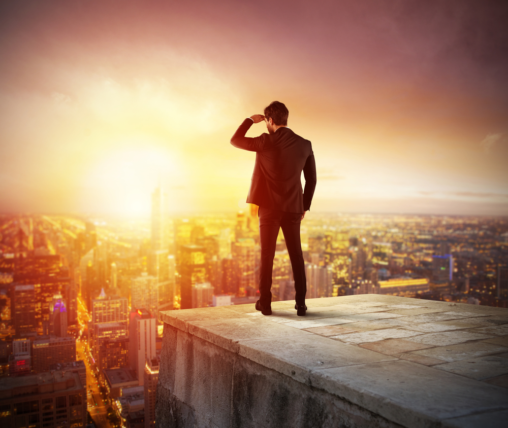 Man in suit standing on rooftop at sunset, looking over a cityscape, with his hand on forehead shielding his eyes.