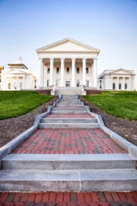 Stone steps leading up to a grand white building with columns and pediment, flanked by two smaller structures housing it services under a clear sky.