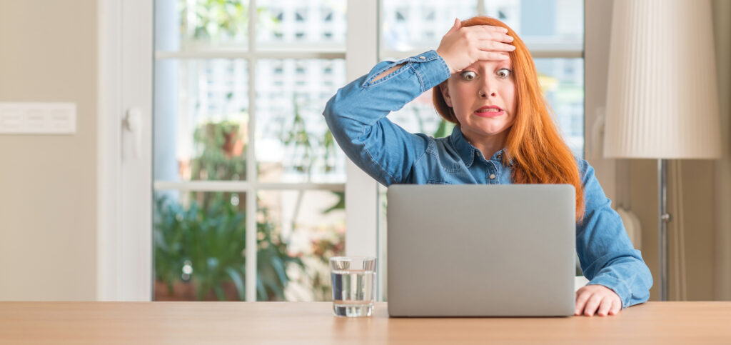 Redhead woman using laptop at home stressed, shocked with shame and surprise for causing a cyber incident.