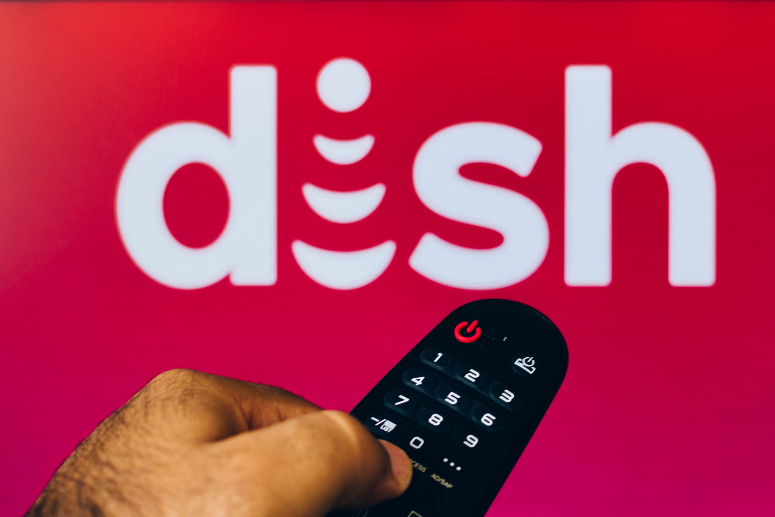 February 21, 2022, Brazil. In this photo illustration a close-up of a hand holding a TV remote control seen displayed in front of the Dish Network Corporation logo