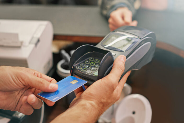 A person uses a blue credit card on a payment processing terminal held by another person at a store checkout.