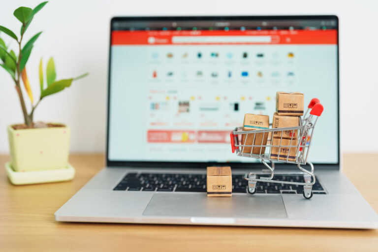A miniature shopping cart filled with small boxes sits on a laptop keyboard, displaying an ecommerce site on the screen, with a potted plant beside it.